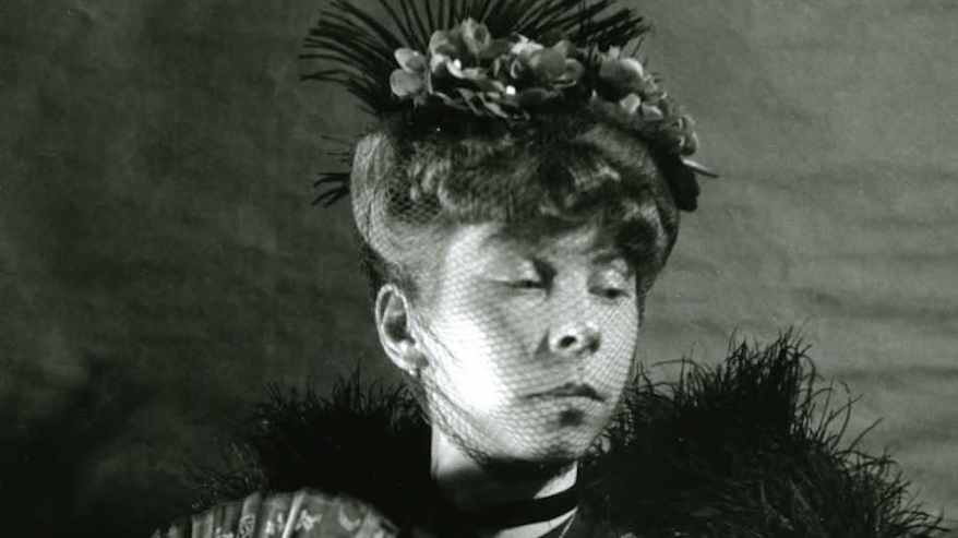 Mustavalkoisessa vanhassa valokuvassa näyttävään asuun pukeutunut nuori nainen, päässä koriste, kädessä viuhka ja pitsikäsineet.