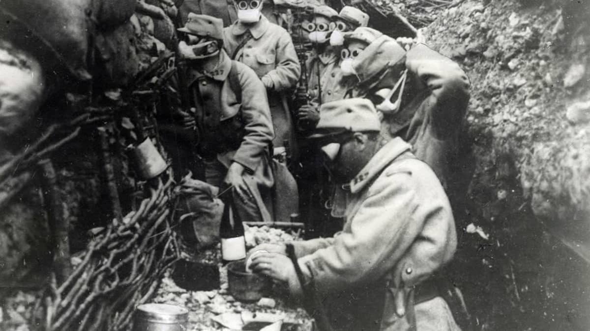 Mustavalkoinen kuva kaasunaamioihin pukeutuneista ranskalaisista sotilaista ensimmäisen maailmansodan juoksuhaudassa. Yksi sotilaista valmistaa ruokaa. 