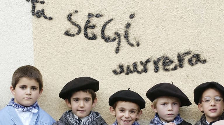 Viisi pikkupoikaa rivissä. Kaikilla on sinikuvioinen kaulaliina, neljällä musta baskeri. Takana seinässä on graffiti, jossa lukee Ta segi aurrera".