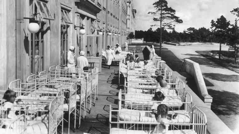 Mustavalkoinen kuva lapsista sängyissä sairaalan terassilla, taustalla valkopukuisia ja -hilkaisia hoitajia. 
