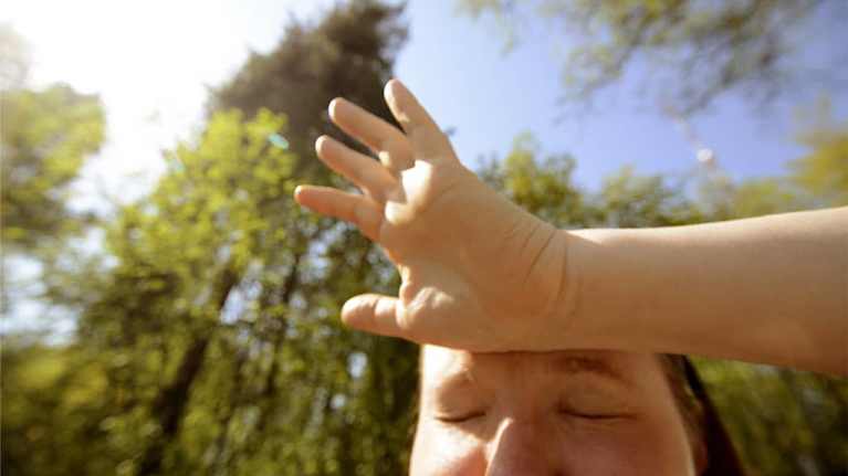 A woman wipes sweat from her forehead against a backdrop of tall fir trees.