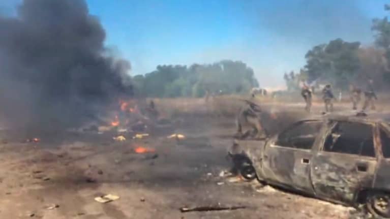 Hiekkakentällä tuhtoutunut autonraato seka jokin palava ja savuava jäännös. Taustalla muutamia sotilaita ja puita.. 