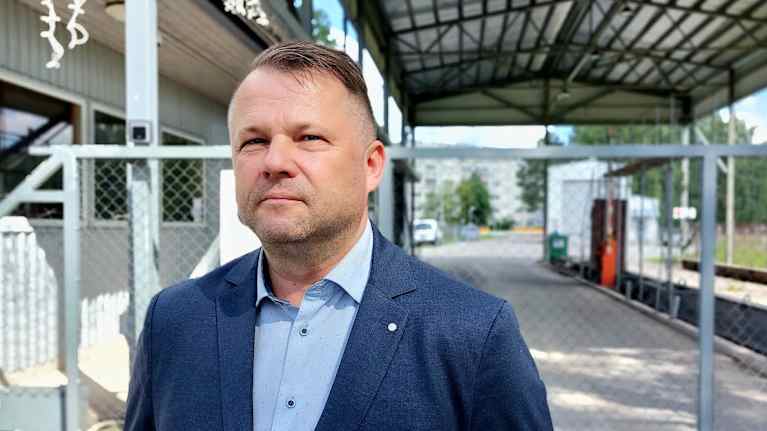 En man i blå kavaj står utomhus framför ett staket och en byggnad med tak i bakgrunden.