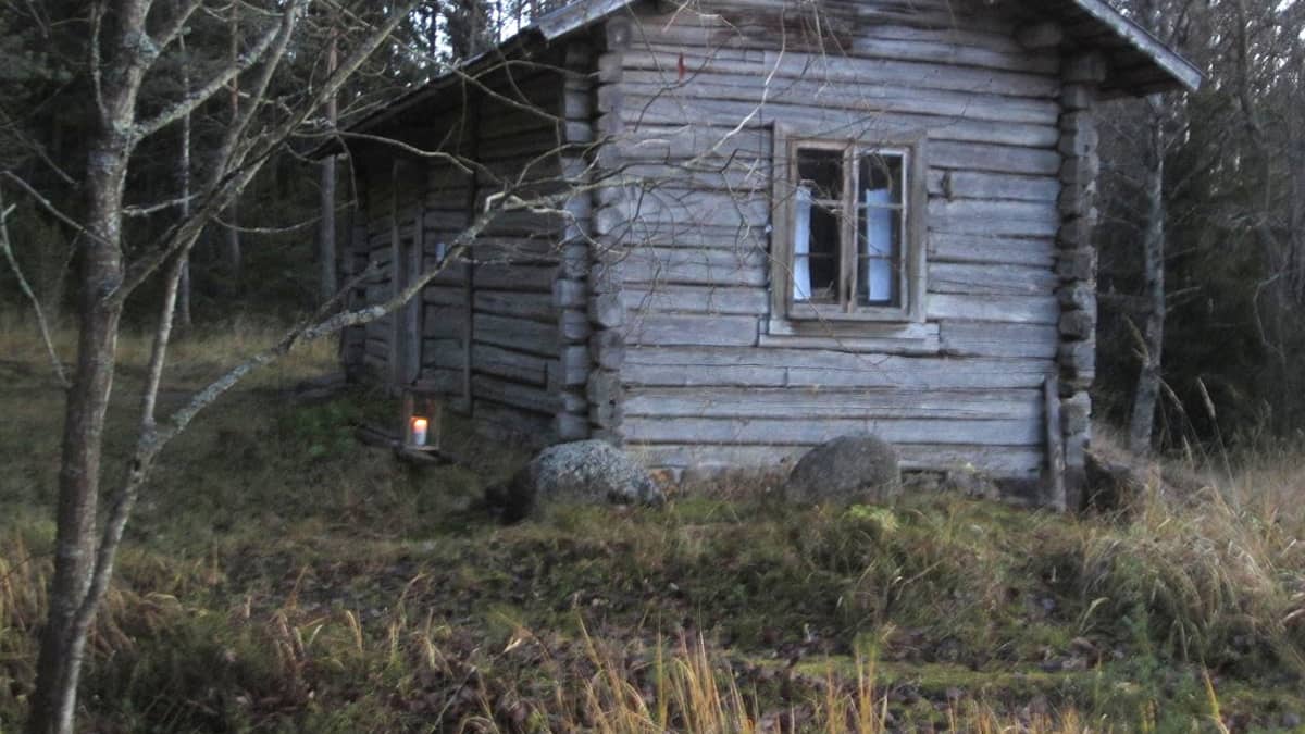 Harmaan, vanhan rakennuksen vieressä palaa kynttilä. Talo on Laitilan Kaariaisten kylän mäkitupa.