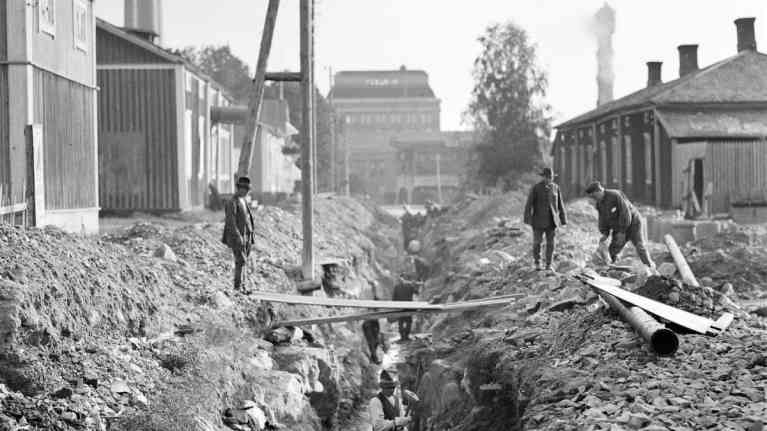Mustavalkoinen kuva miehistä kaivamassa kadulla ojaa viemäriputkille. Taustalla tehdasrakennus ja savua tupruttava tehtaanpiippu.