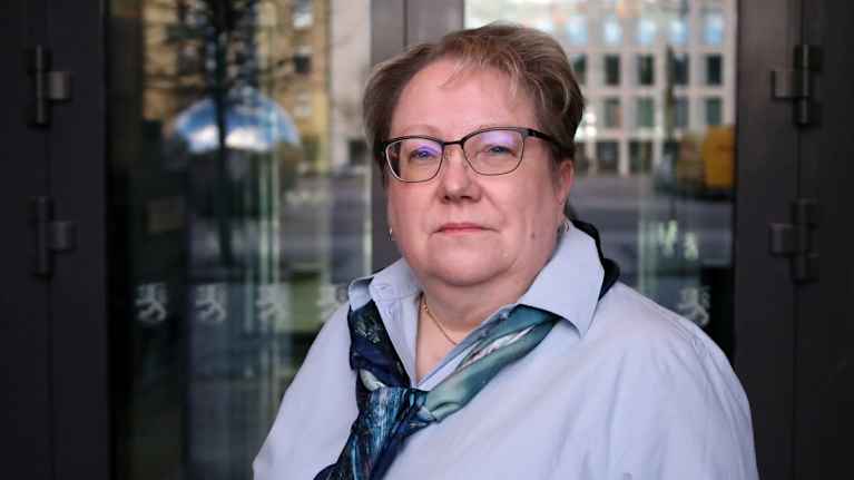 A woman with short hair, glasses, a blue shirt and a scarf looks seriously at the camera.