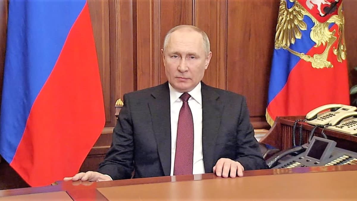 Venäjän presidentti istuu pöydän takana tummassa puvussa ja viininpunaisessa kravatissa. Taustalla näkyy kaksi Venäjän lippua seinää vasten.