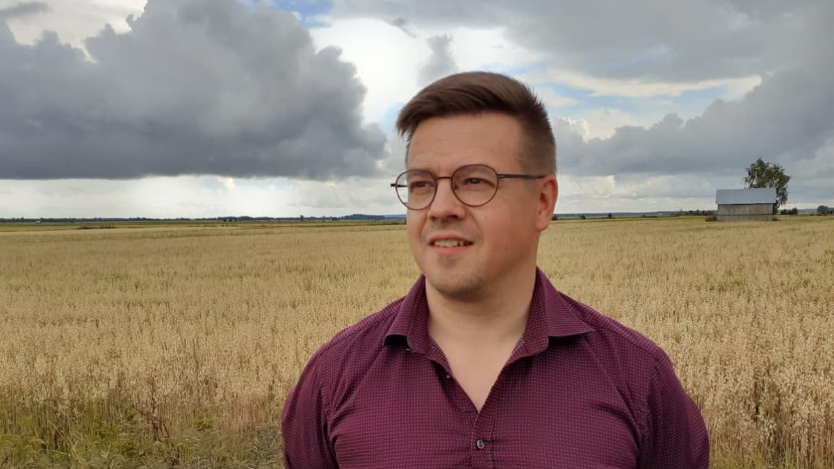 Silmälasipäinen mies seisomassa pellolla eteläpohjalaisen lakeuden keskellä, taustalla heinälato.