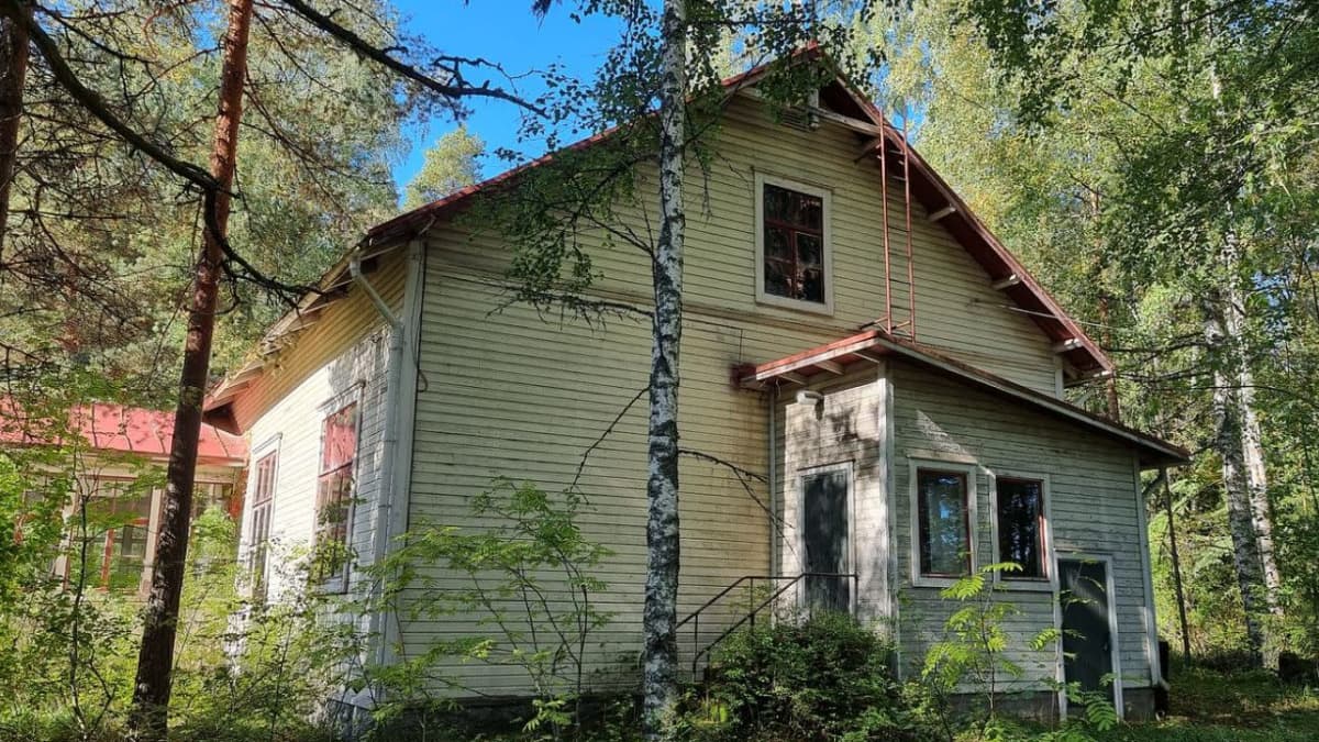 Vaalea puinen rakennus, jossa on punainen harjakatto. Rakennuksen ympärillä on metsää.