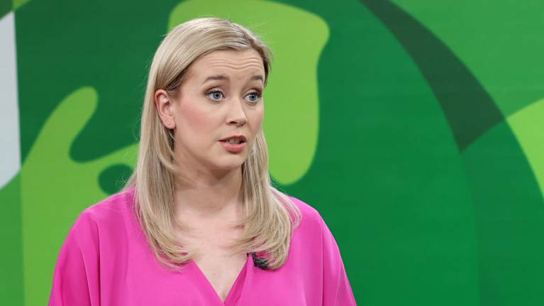 Woman with shoulder-length hair and wearing a pink blouse talking, with a green patterned background.