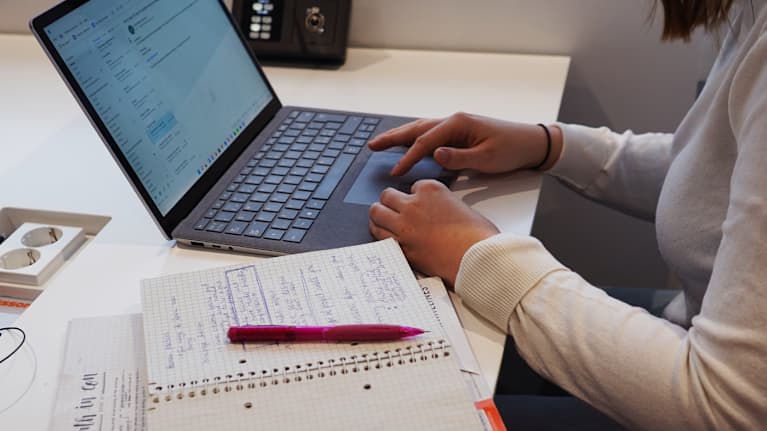 En person som sitter och jobbar med en bärbar dator. Fingarna rör datorns "touchpad" och bredvid personen syns ett anteckningsblock en kulspetspenna.