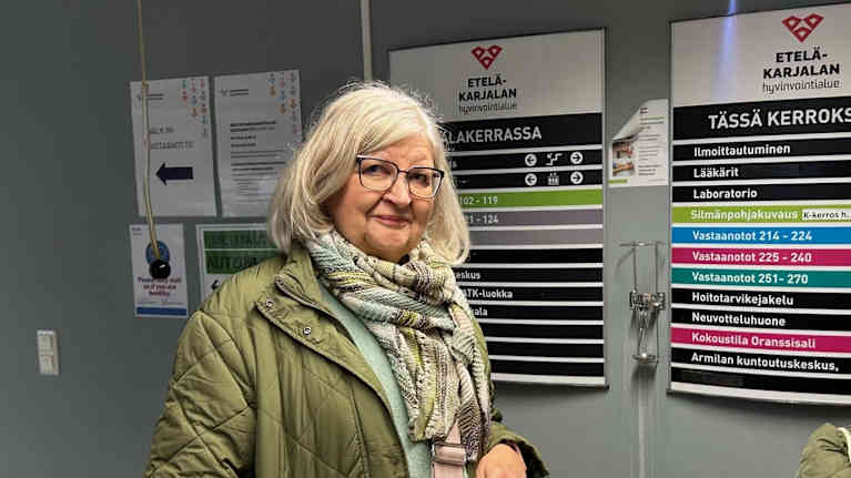 Lähikuva vaaleahiuksisesta eläkeläisnaisesta. Taustalla opaste, jossa hyvinvointiaseman palvelupaikkoja.