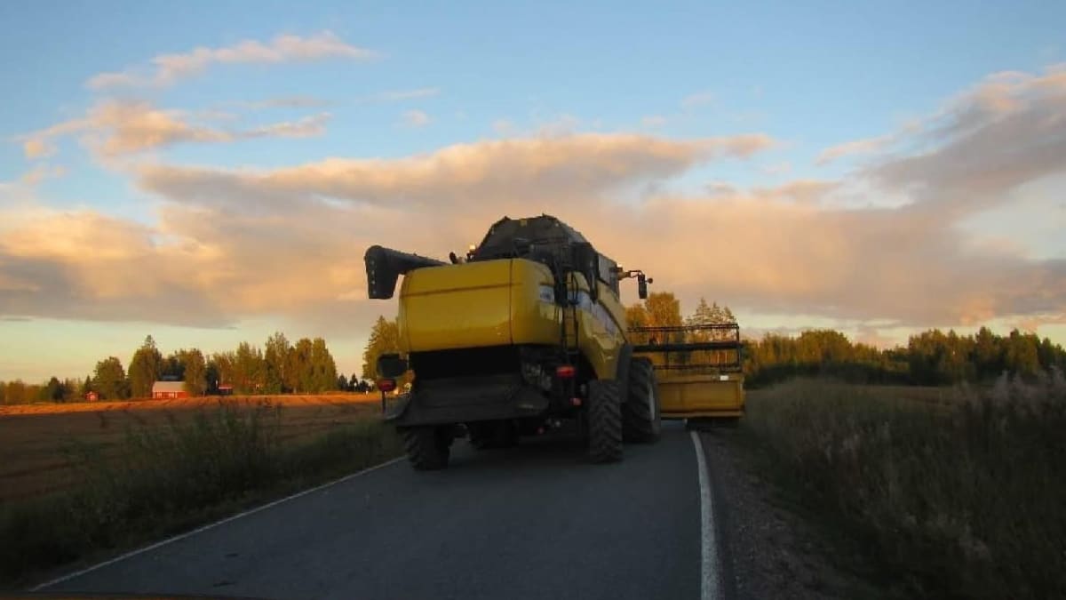 Keltainen  leikkuupuimuri peltojen keskellä menevällä tiellä. Kuvan etualalla näkyy henkilöauton etuosaa. Pilvinen iltataivas. 