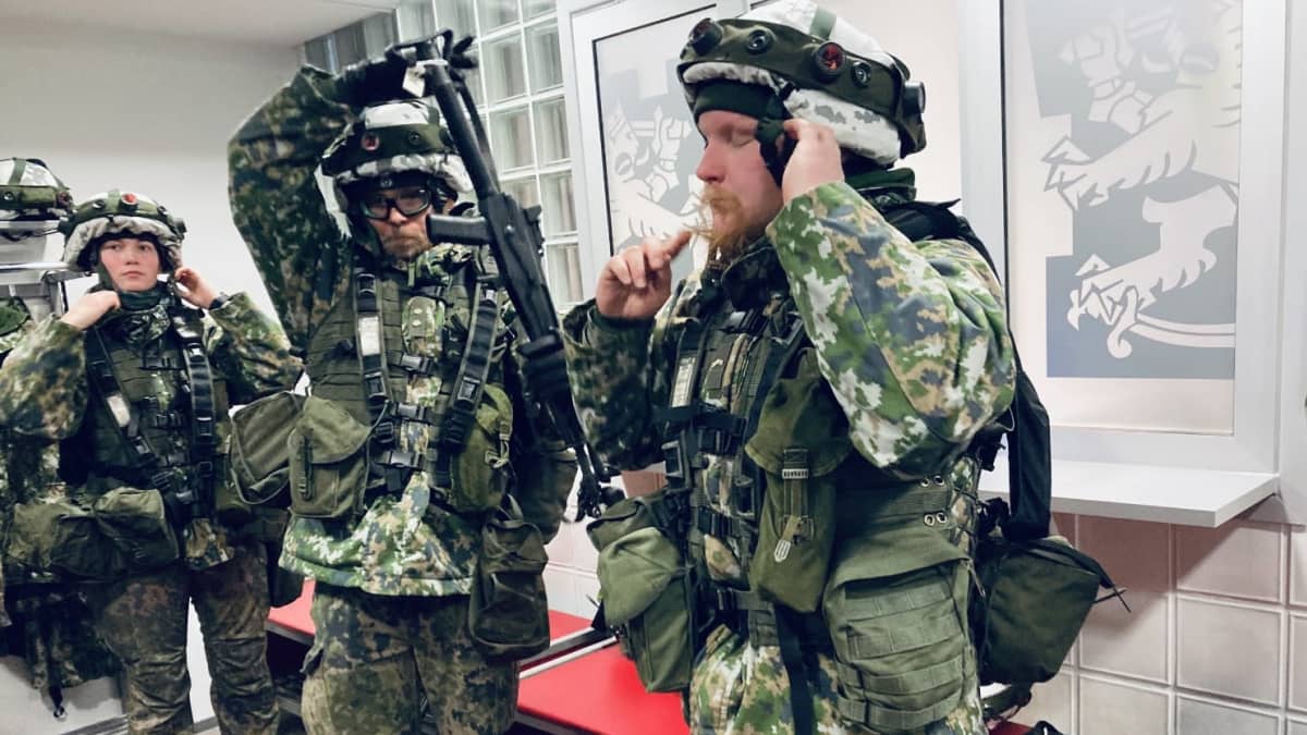 Kolme maastopukuihin pukeutunutta reserviläistä pukee taisteluvarustusta päälle aulatilassa.