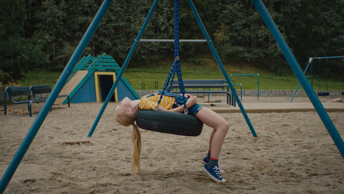 Vaaleahiuksinen tyttö makaa rengaskeinussa selällään keskellä leikkipihaa.