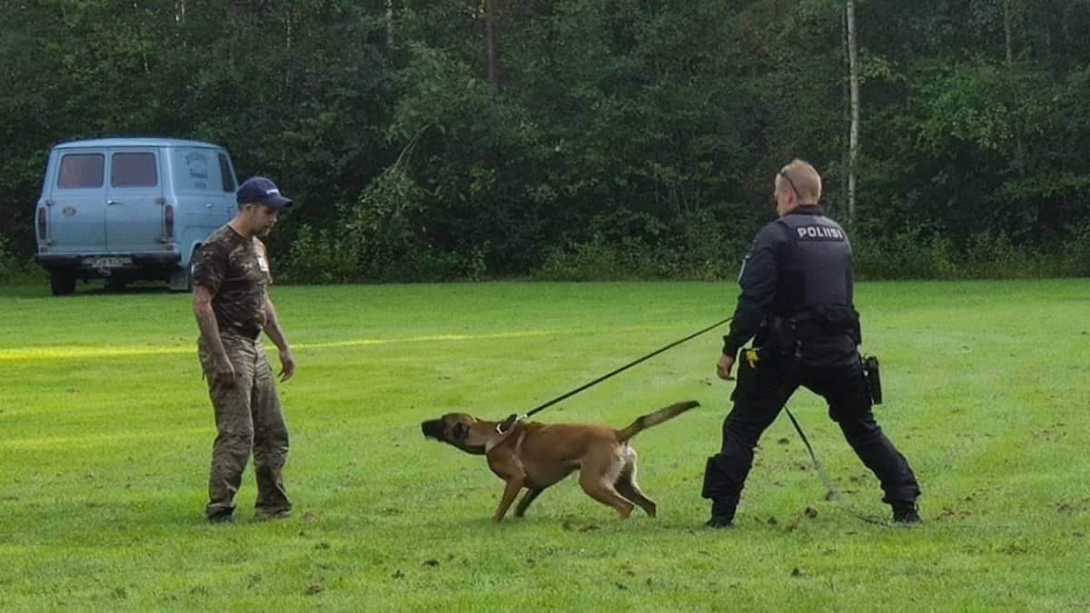En polis håller en polishund i ett koppel. Framför hunden står en man.