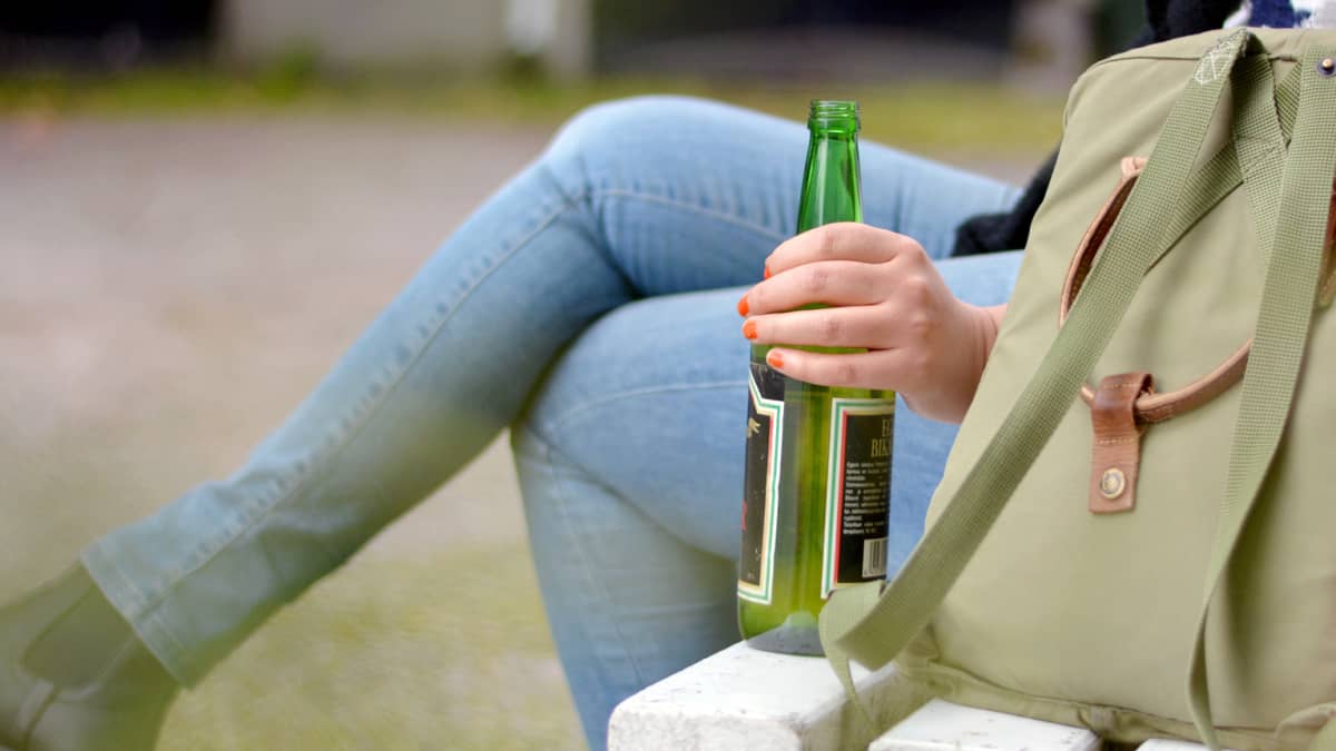 En person sitter på en bänk med en ryggsäck och vinflaska bredvid sig.
