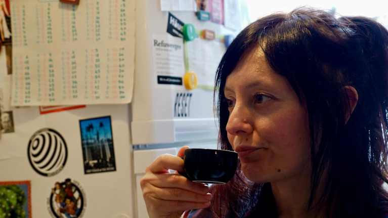 Lähikuvassa kahvia juova tummatukkainen nainen, taustalla näkyy jääkaappiin magneeteilla kirjoitettuja lappuja ja kuvia.