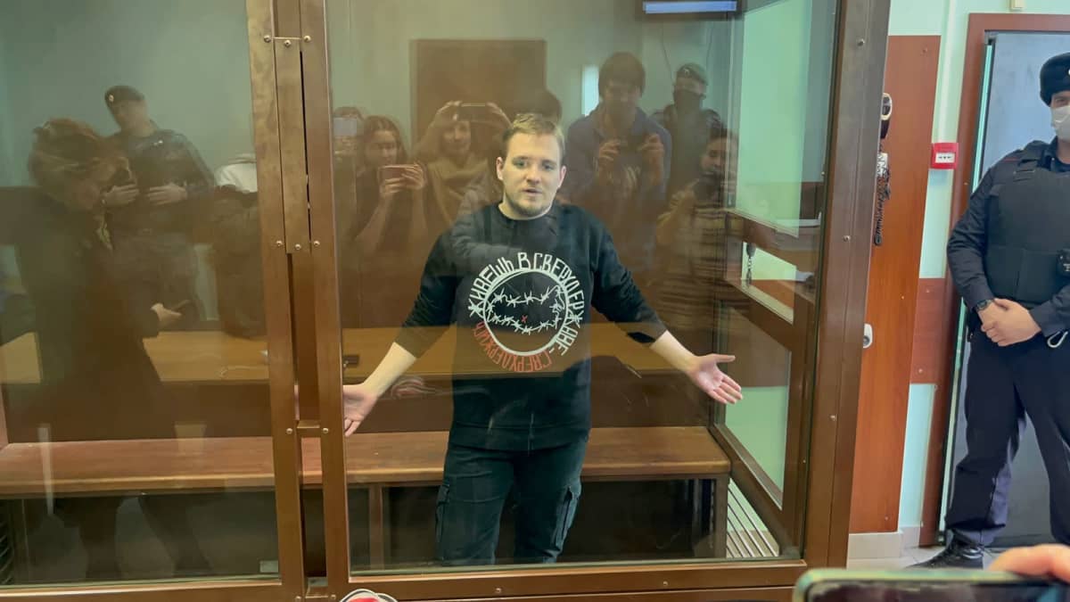Dmitri Ivanov seisoo oikeussalissa lasiseinäisessä kopissa ja levittelee käsiään. Hänellä on parta, musta paita ja tummat housut. Kopin vieressä seisoo vartija. Lasiseinästä heijastuvat Ivanovia kuvaavat ihmiset.