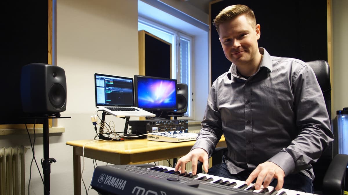 Nuori mies soittaa kosketinsoitinta ja hymyilee kameralle studiomaisessa huoneessa.