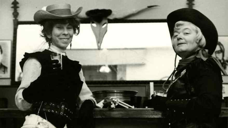 Kaksi henkilöä cowboy-asuissa hymyilee kameralle, toisella lasi kädessä, naamiaisjuhlat.