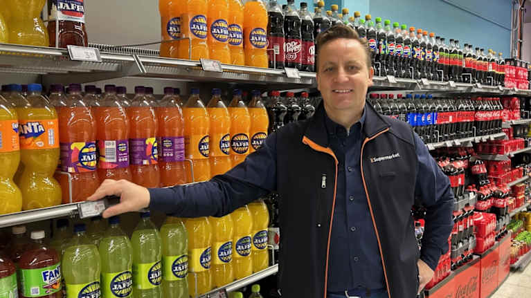 Henkilö on ruokakaupan juomaosastolla, jossa on tarjolla erilaisia virvoitusjuomia. Hän nojaa hyllyyn.