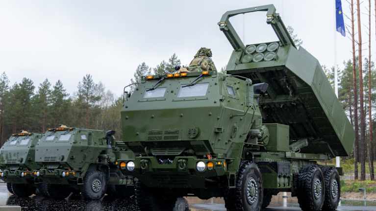 Three large, dark green military trucks in a parking lot, one vehicle is outfitted with a rocket launching system on the back which is pointed upward.