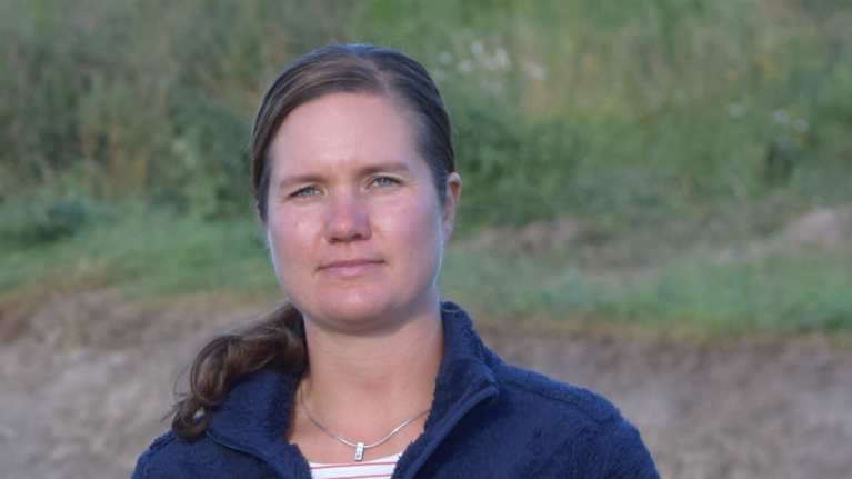 En person står på en åker eller äng, i bakgrunden syns en liten bassäng med stenkarnter, grönt gräs och åker, skog.