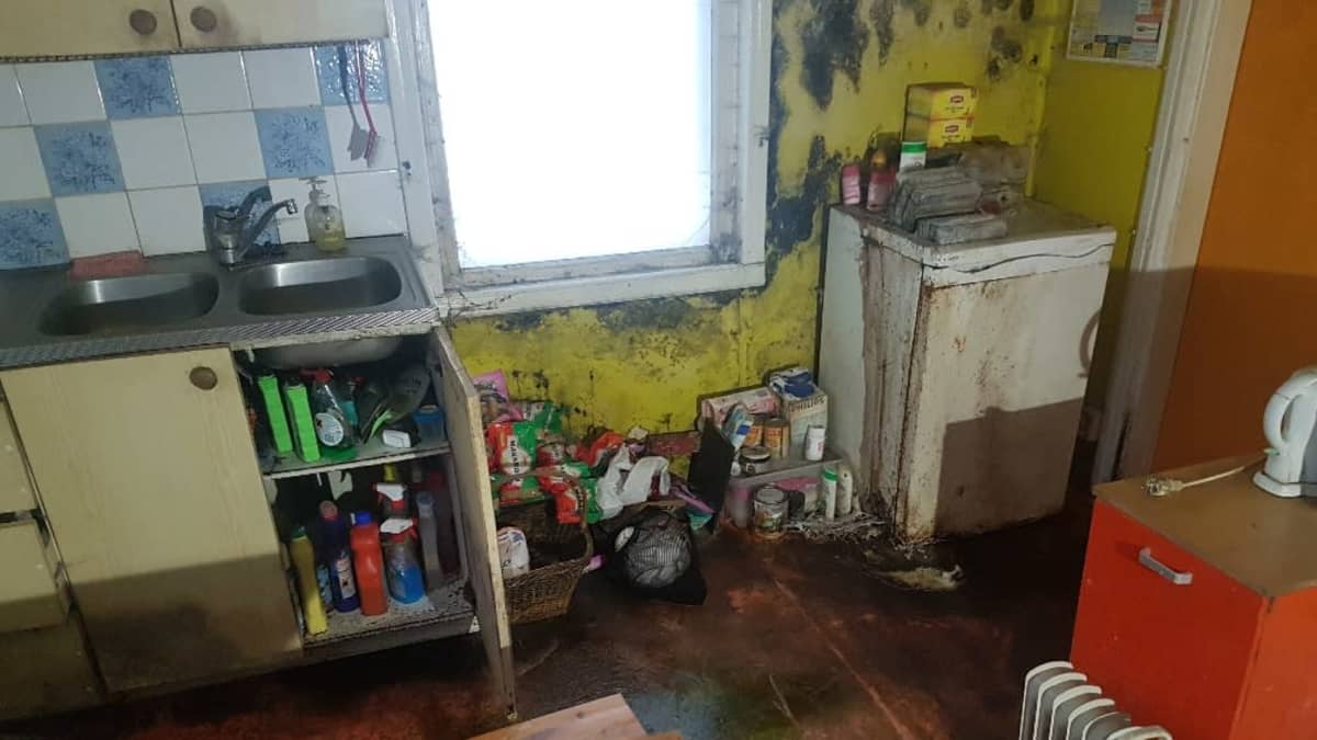 Kuva tyhjilleen jätetyn omakotitalon keittiöstä. Keltaisilla seinillä on mustia jälkiä, ja myös lattia ja ruokapöytä ovat tummuneet. Lattialla on elintarvikkeita, kuten makaronipusseja.