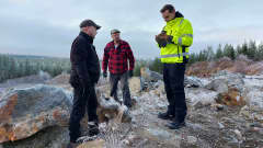 Kolme miestä tutkii kivinäytettä kallioisessa maastossa.