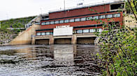 Punatiilinen vesivoimalaitos iijoella, etualalla vettä ja rantapusikkoa.