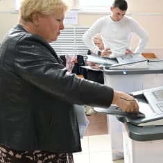 Vaaleahiuksinen nainen mustassa takissa syöttää äänestyslippua sähköiselle äänestyslaitteelle. Taustalla nuori mies valkoisessa paidassa tarkastelee vastaavaa konetta kädet lanteillaan. Seinällä on vaalijuliste.