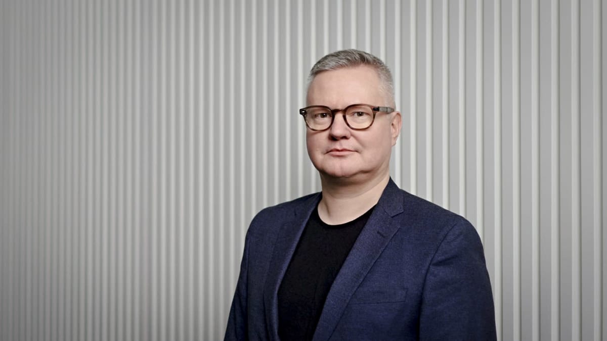 Janne Hukkanen seisoo vaalean seinän edessä, katsoo kameraan puvun takki yllään, puolikuva ja neutraali ilme.