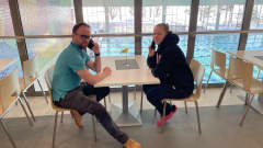 Pöydän ääressä istuvat mies ja nainen puhuvat puhelimessa. Taustalla näkyy uimahallin uima-allas.