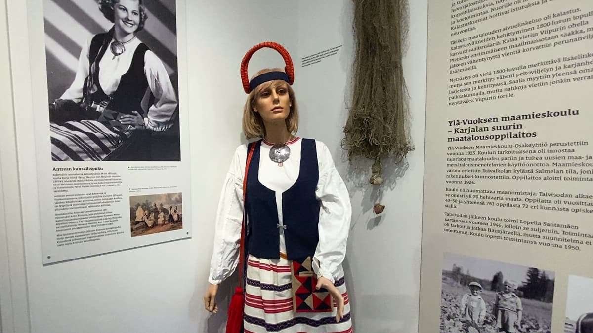 Kansallispukuun puettu mallinukke seisoo Armi Kuuselaa esittään kuvasuurennoksen vieressä.