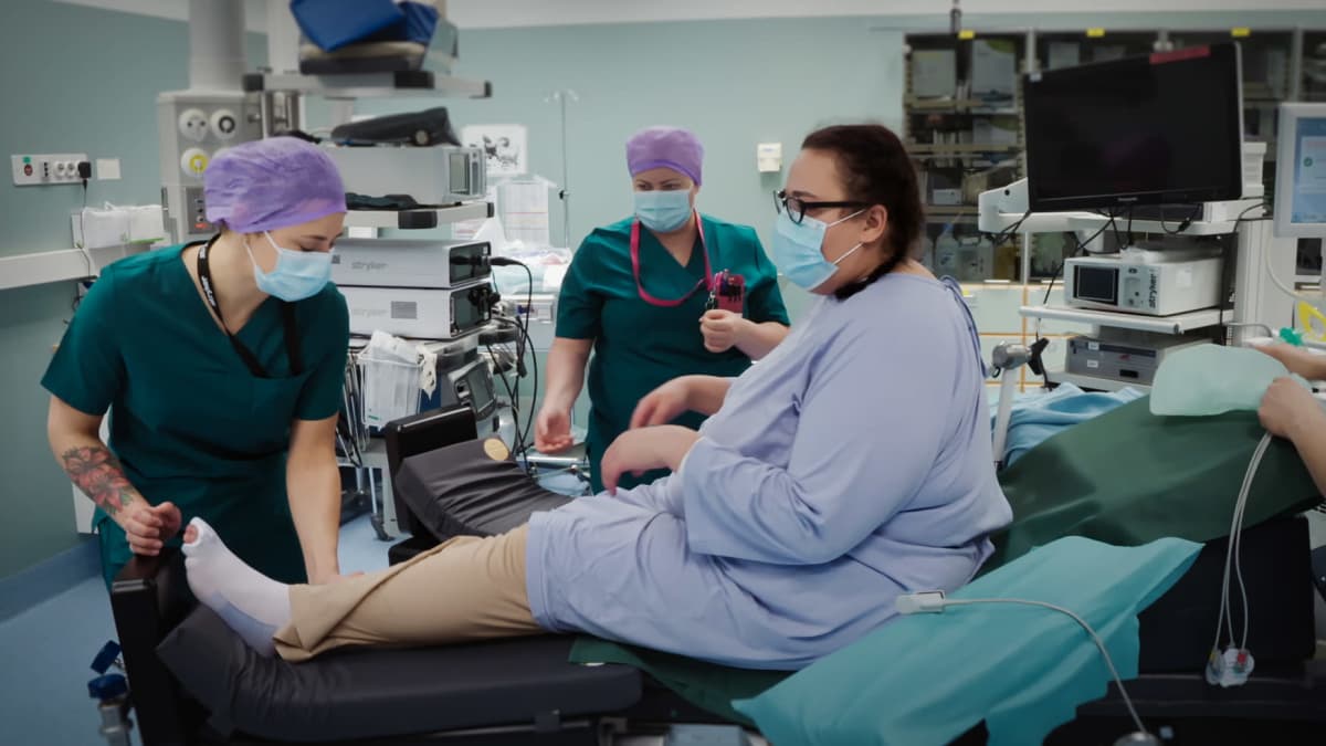 Sofia Heinonen istuu leikkaussalissa leikkauspöydällä, hoitaja asettelee hänen jalkaansa ylös. Toinen hoitaja taustalla.
