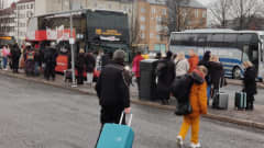 Busseja on pysäkeillä. Ihmiset odottavat nousua busseihin. Monella on mukanaan matkalaukkuja. 