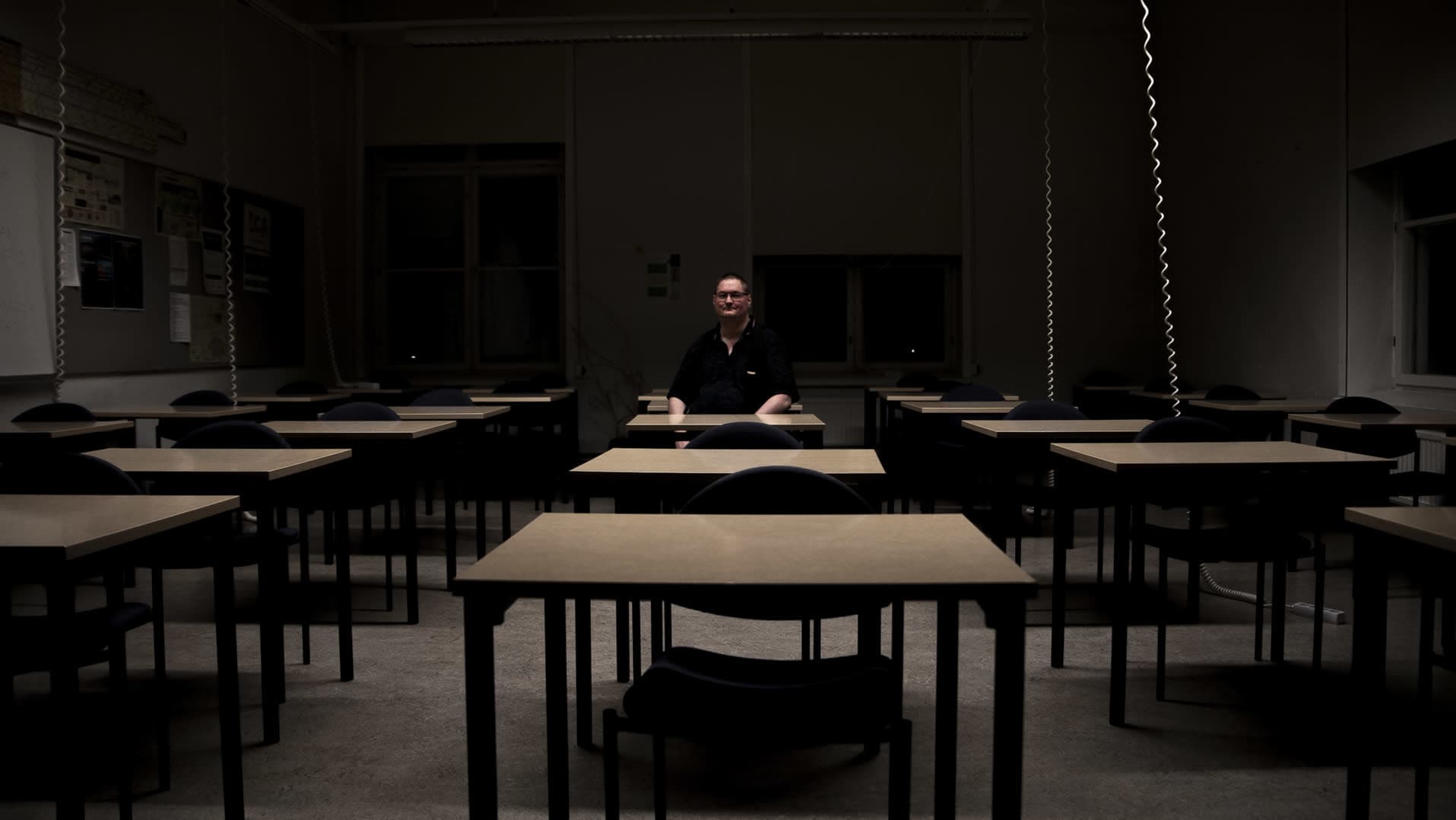 Mies istuu valokeilassa yksin pimeässä luokkahuoneessa.