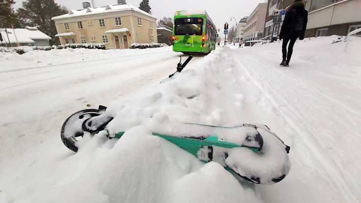 A green e-scooter on the ground in a snowbank, alongside a snowy street and sidewalk.