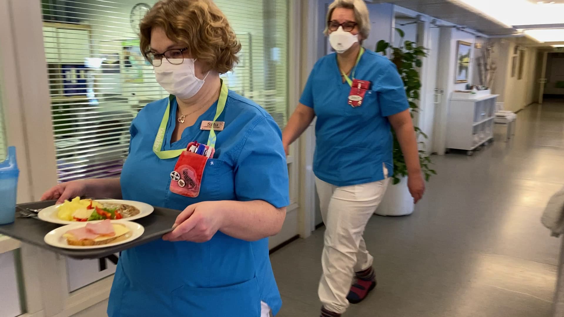 Kaksi siniasuista hoitajaa käytävällä vie ruoka-annoksia potilaille 