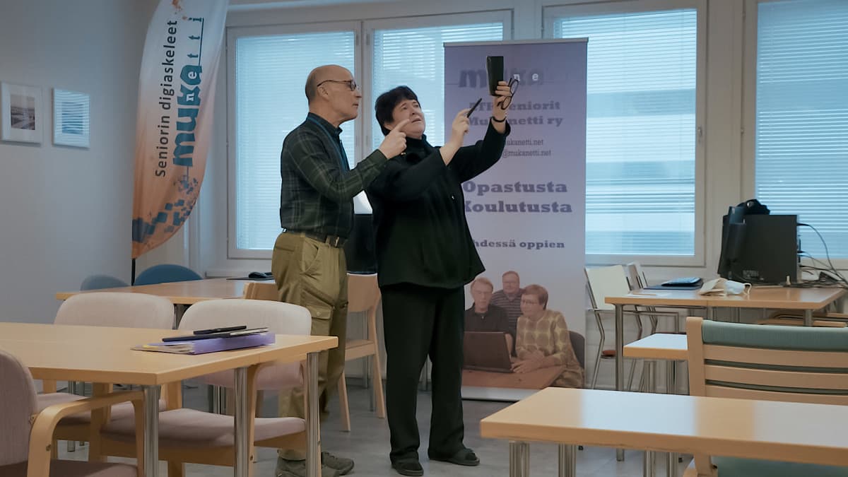Seniorimies ja seniorinainen seisovat luokkatilassa. Nainen on nostanut älypuhelimen toisella kädellään ylös. Mies osoittaa älypuhelinta.
