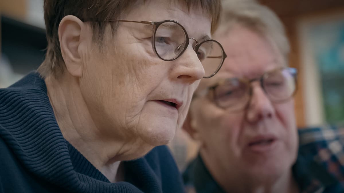 Etualalla lähikuvassa silmälasipainen seniorinainen. Taustalla epätarkkana harmaapäinen seniorituutorimies.