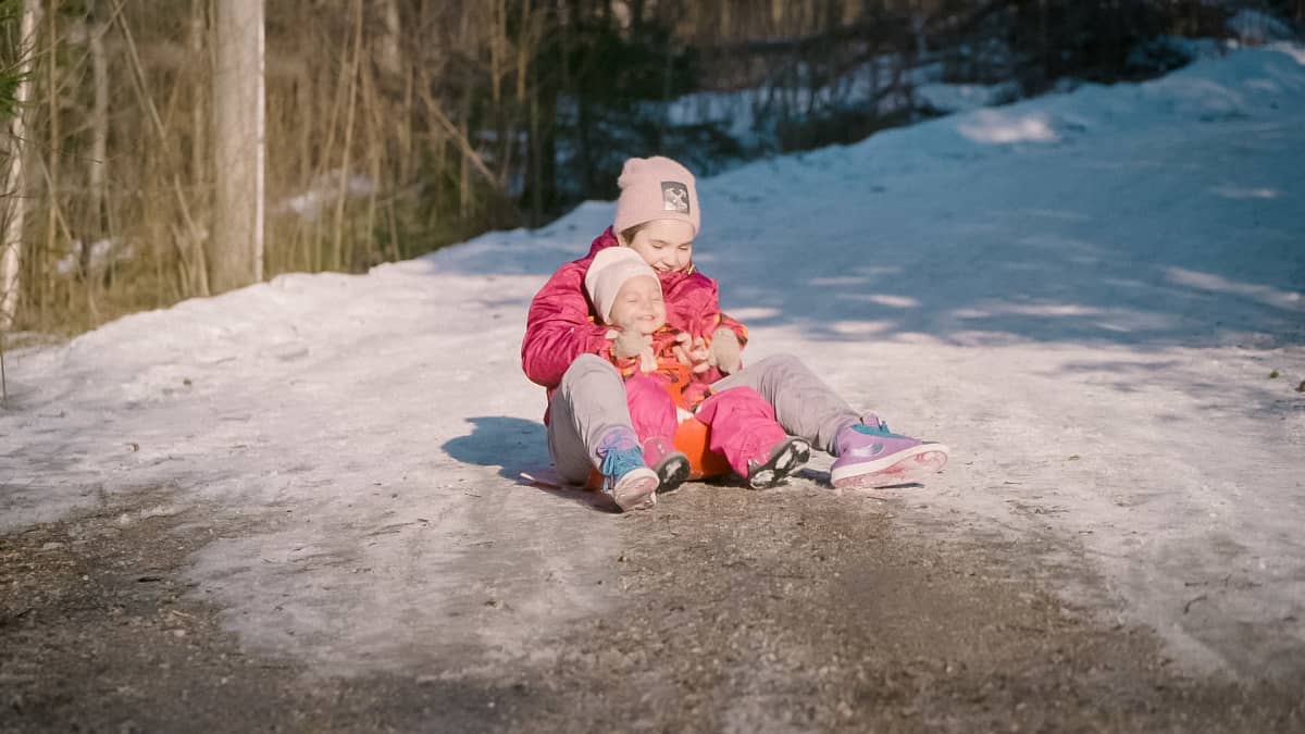 Isompi lapsi laskee pulkkamäkeä taaperoikäinen lapsi haarojensa välissä. Lasku päättyy, kun lumi vaihtuu hiekaksi, ja lapsia naurattaa.