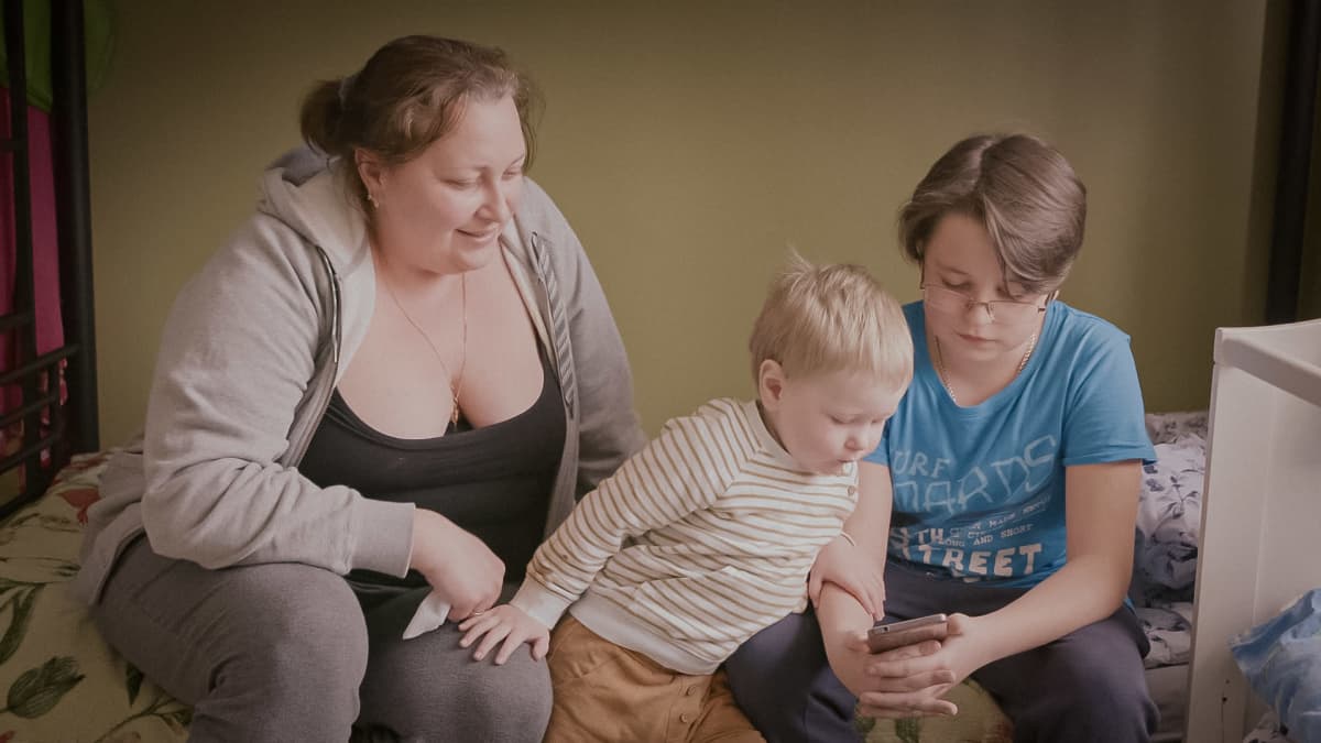 Vovan äiti, Vovan pikkuveli ja Vova soittavat isälle videopuhelua istuen sängyn laidalla.