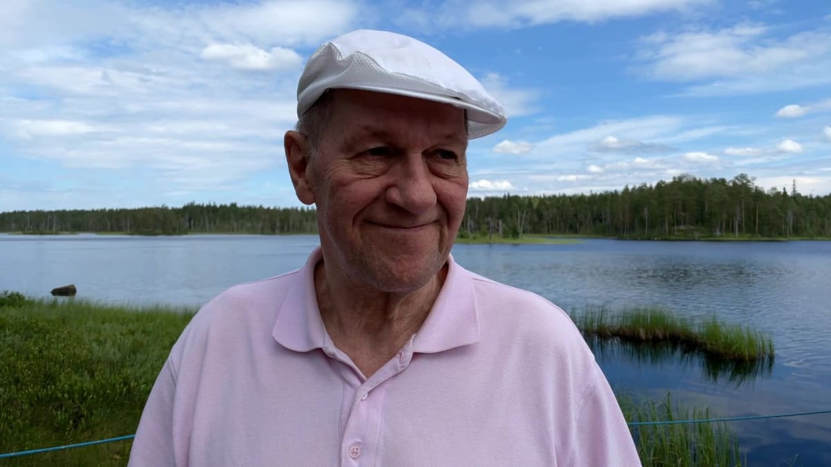 Vanha mies seisoo valkoinen lippalakki päässä järven rannalla.