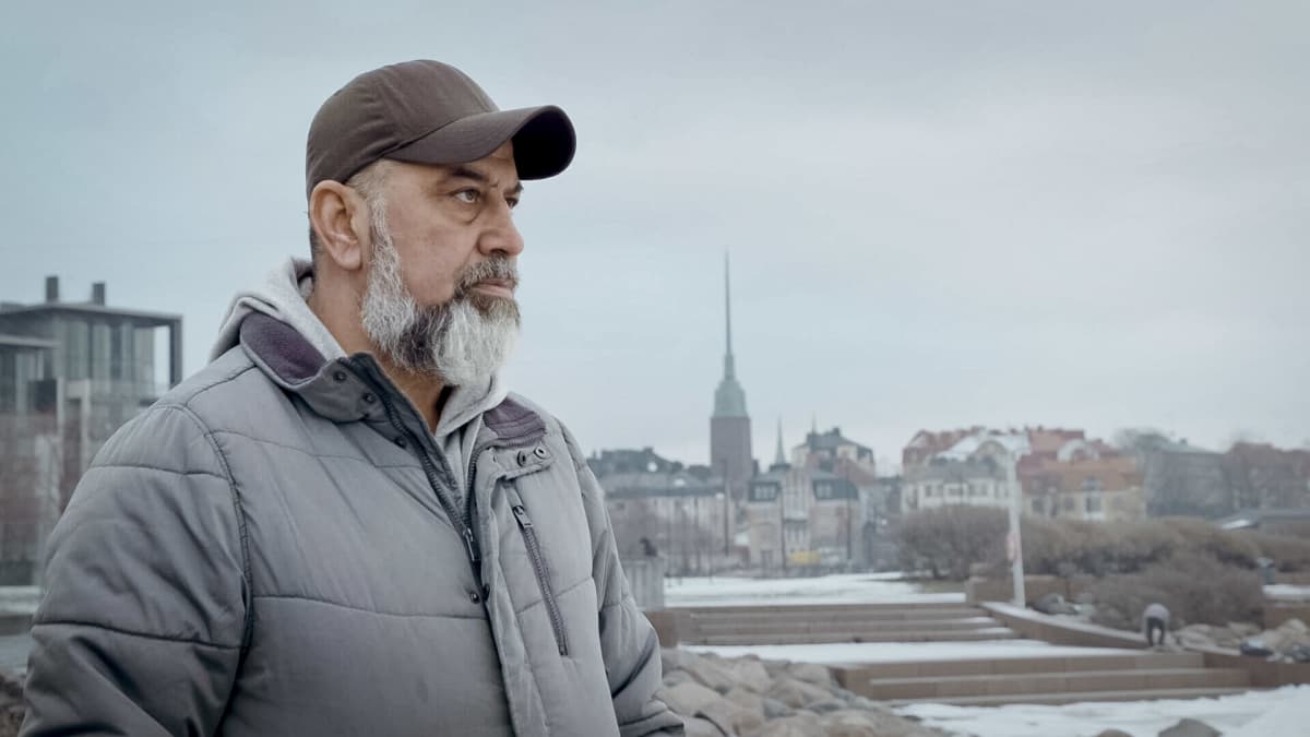Keski-ikäinen parrakas mies katsoo lippalakki päässä eteensä, taustalla Helsingin kaupunkia.