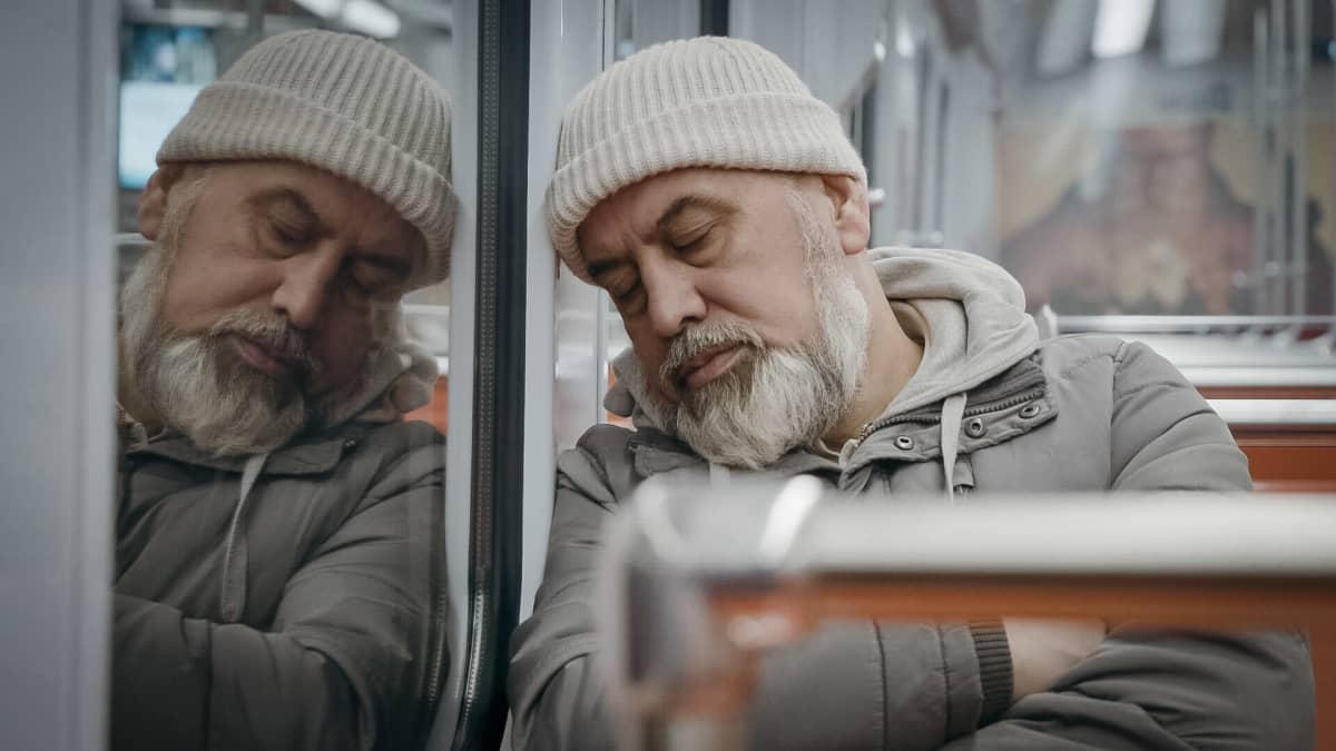 Keski-ikäinen mies istuu metrossa ja nojaa silmät kiinni ikkunaan.