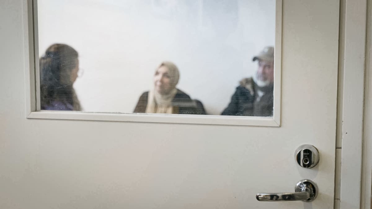 Ovessa olevan lasin läpi näkyy kolme sumeaa hahmoa istumassa huoneessa.