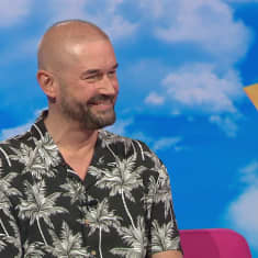 Ohjaaja, koreografi Marco Bjurström istuu Puoli seitsemän -ohjelman pinkillä sohvalla ja hymyilee. 