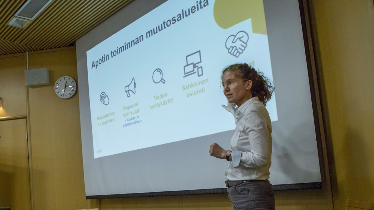En kvinna, Piia Vuorela, står och pekar mot en powerpoint presentation om den nya patientinformationssystemet Apotti. 
